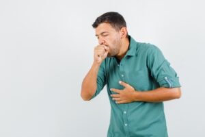 A man wearing a green shirt is coughing and holding his chest with a pained expression.